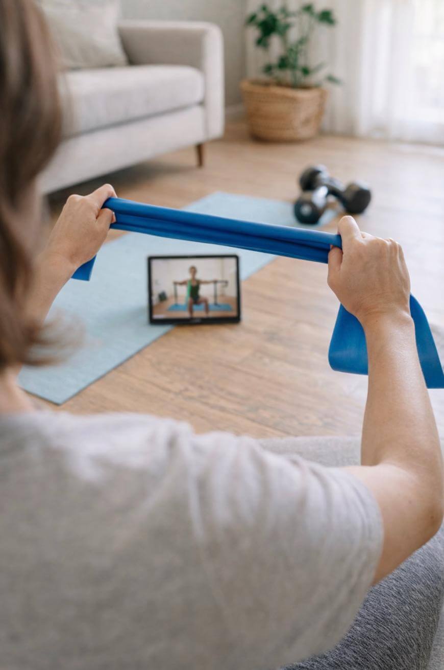 A person holds a blue resistance band whilst following an exercise video on a tablet in a sitting room. A yoga mat, dumbbells, and a potted plant are visible in the background.