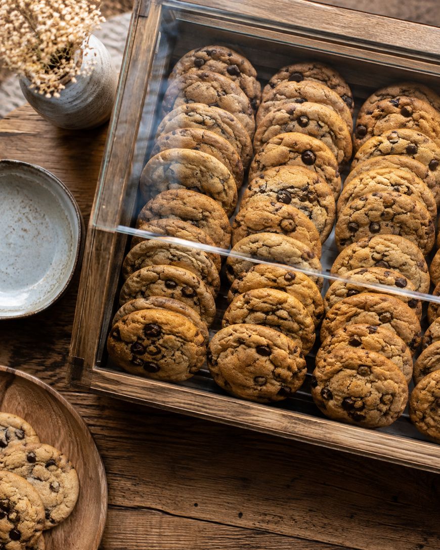 A wooden display case filled with rows of chocolate chip cookies on a rustic table, with empty ceramic plates and a small vase of dried flowers nearby.