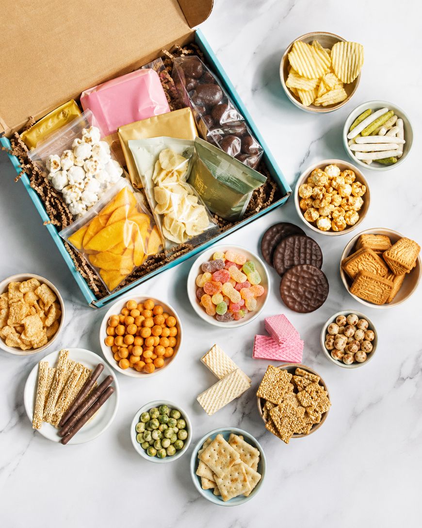 A gift box filled with assorted snacks, including crisps, crackers, sweets, popcorn, wafers, chocolate, and more, displayed in bowls on a white surface around the open box.