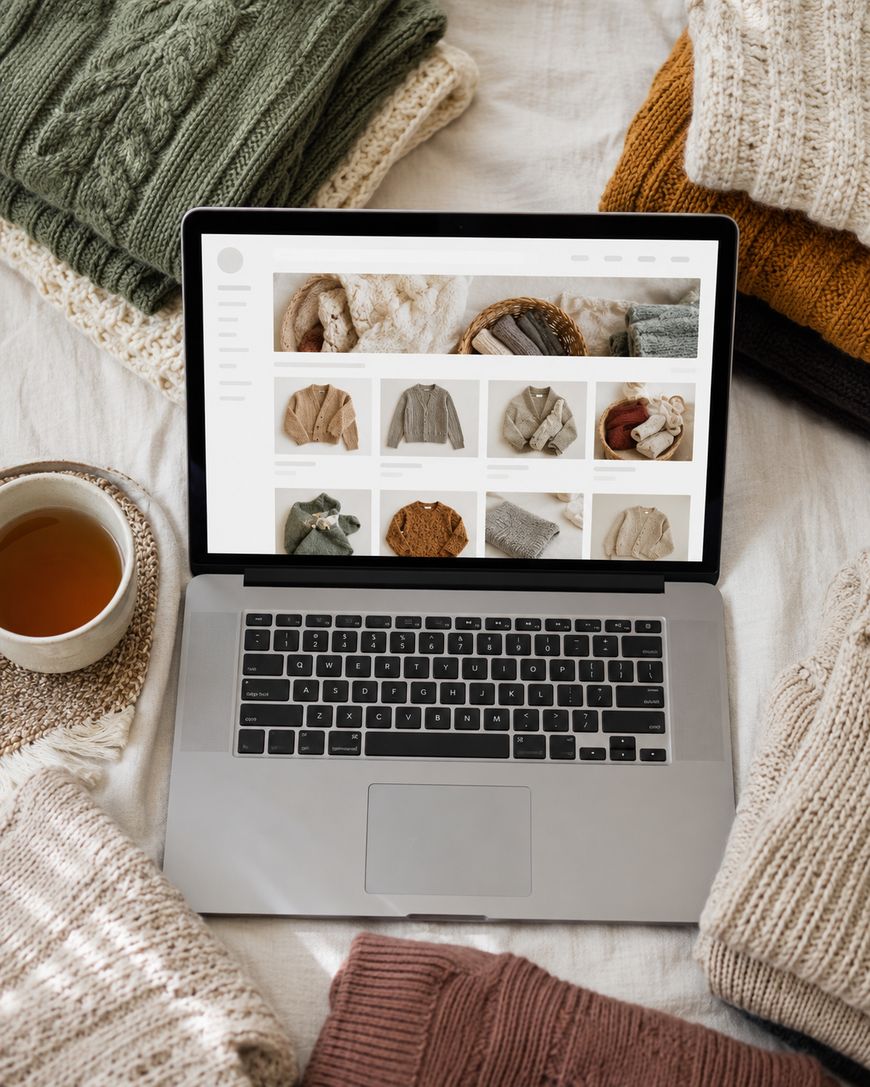 Laptop screen showing an online shop grid of handmade knitted sweaters and cardigans, surrounded by folded knitwear and a cup of tea.