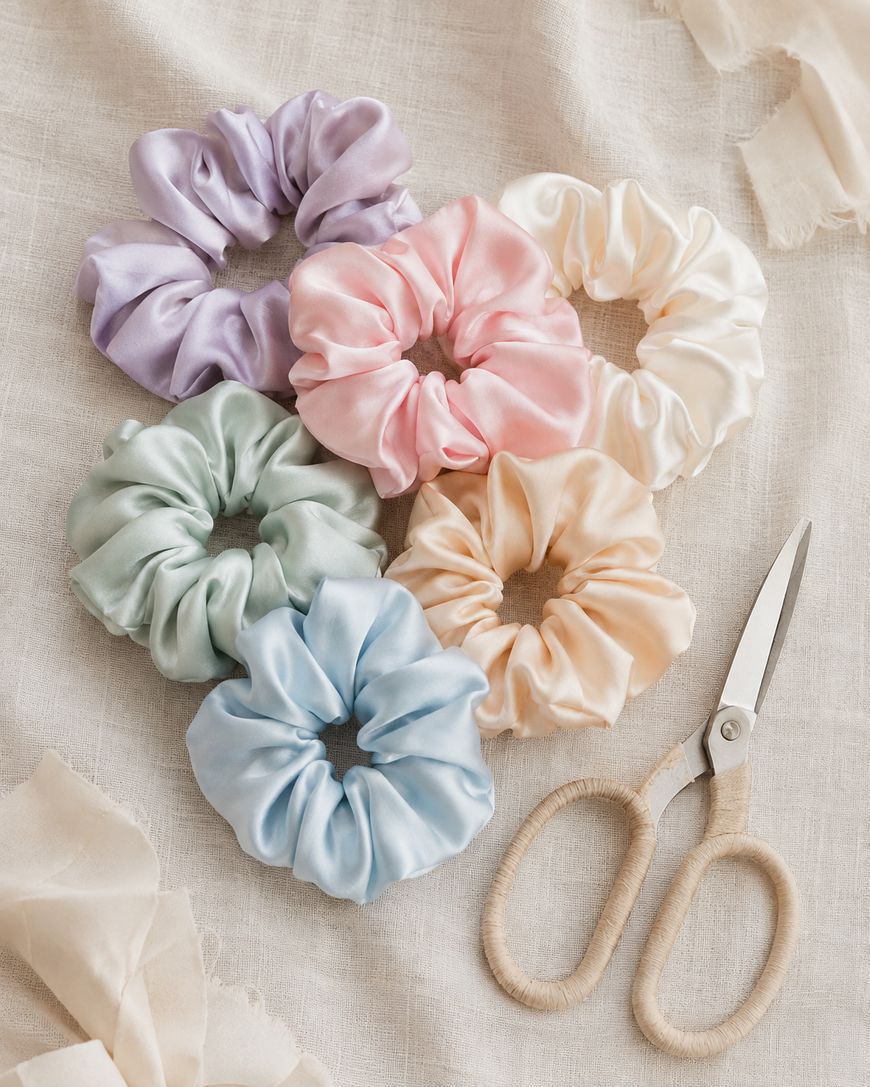 Pastel silk scrunchies in sage, pink and cream arranged on fabric beside a pair of scissors.